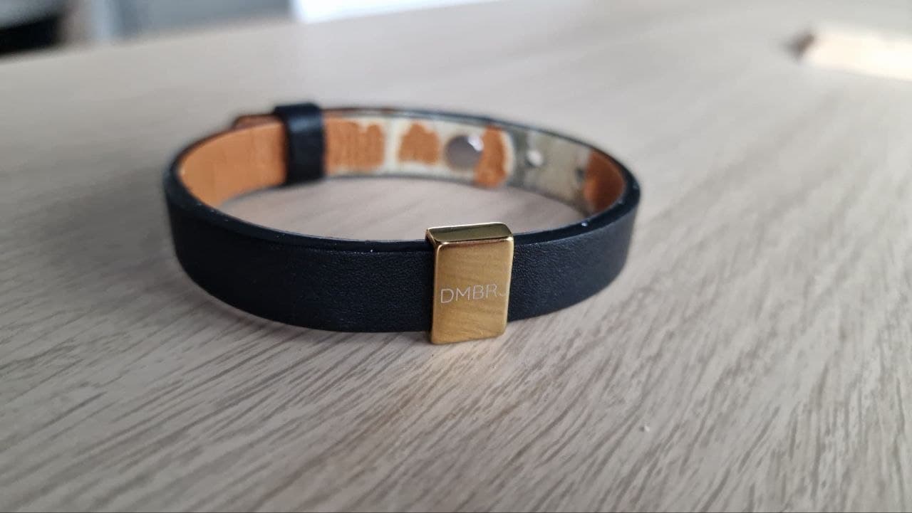 A black leather bracelet with a small gold plate engraved DMBRJ, lying on a wooden table